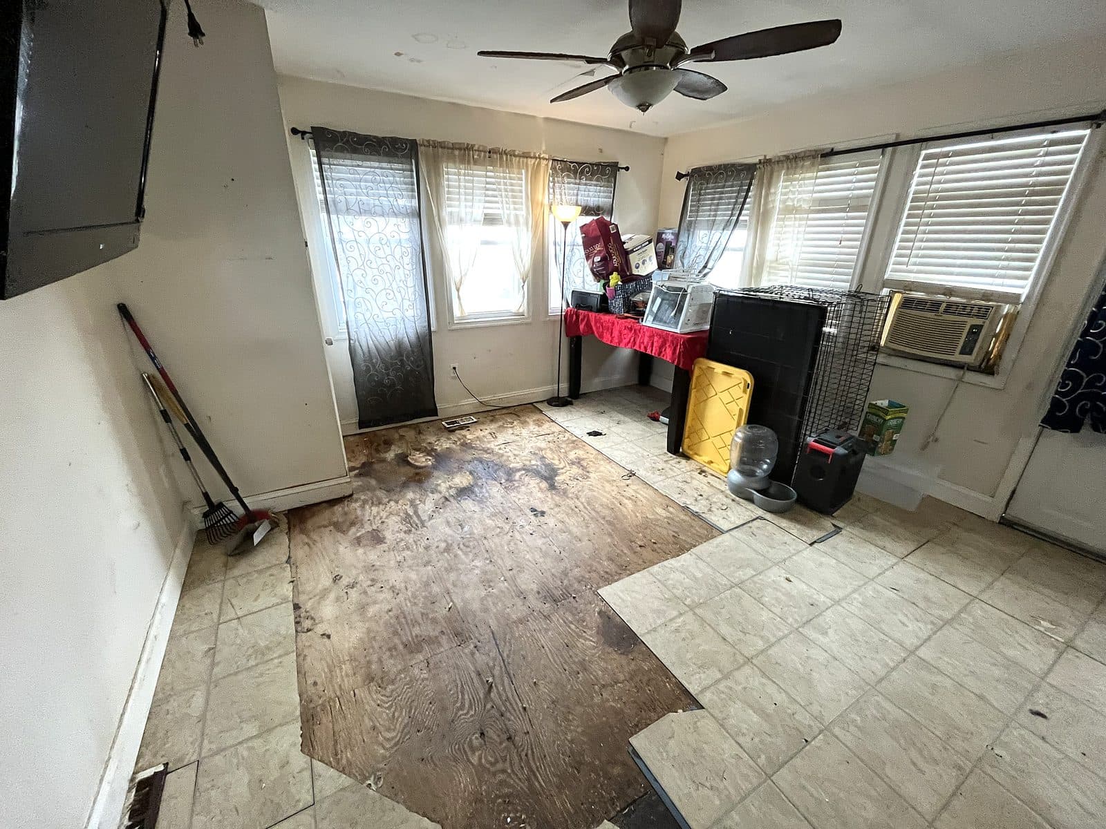 Living room with the floor tile torn up down to stained plywood, a pet crate and belongings still in the room