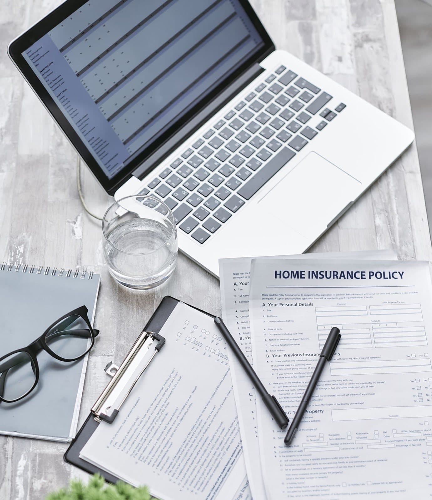 A home insurance policy form on a desk beside a laptop, glasses and pens